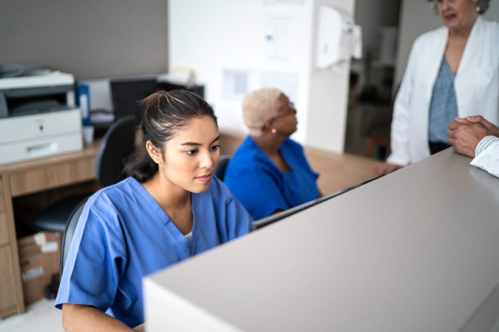 A medical assistant in blue scrubs works at a reception desk computer while a colleague and a patient interact in the background.