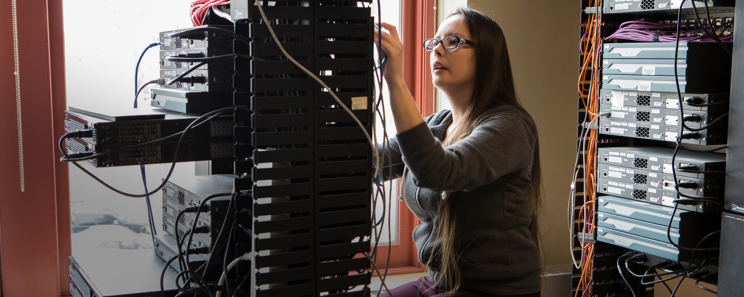 A female student adjusts cables on a server rack in a network lab filled with patch panels, switches, and colourful ethernet cables.