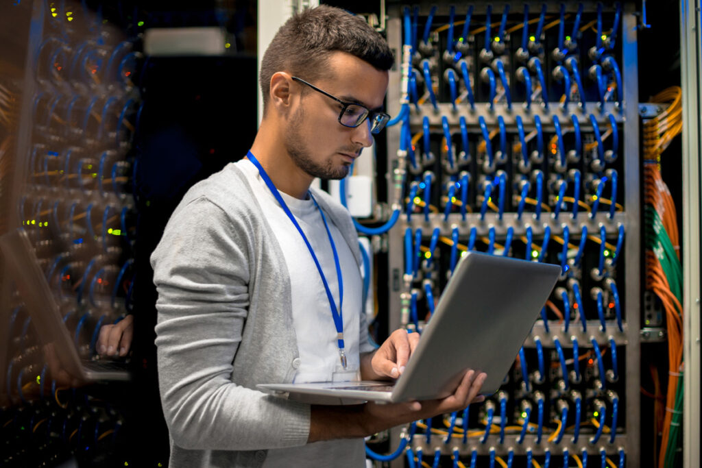 A male IT technician wearing glasses and a lanyard works on a laptop in front of a densely cabled server rack lit with blue and orange indicator lights.