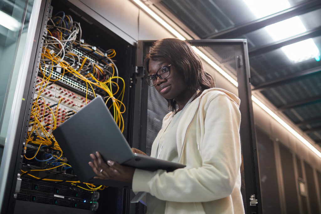 A female network administrator wearing glasses holds a laptop while inspecting an open server rack filled with yellow and orange cables in a data centre.