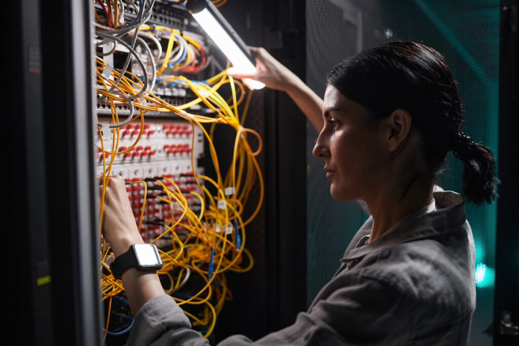 A female network technician with a ponytail traces cables on a patch panel inside a server cabinet illuminated by a work light.