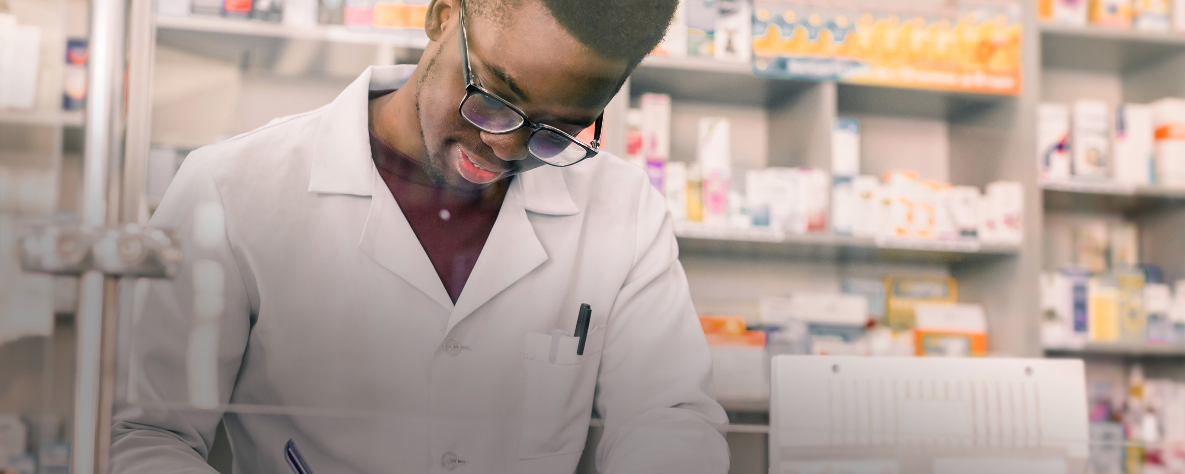 A male pharmacy technician in a white lab coat writes notes at a counter, surrounded by shelves stocked with pharmaceutical products.