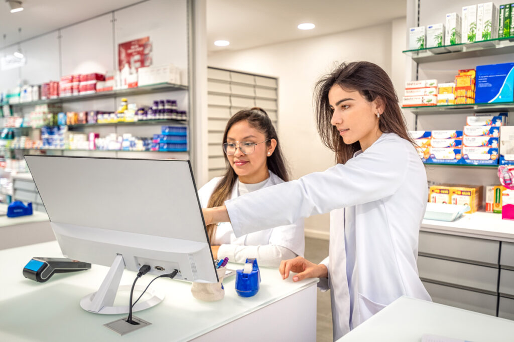 Two pharmacy technicians in white lab coats collaborate at a computer terminal behind a pharmacy counter stocked with medication products.