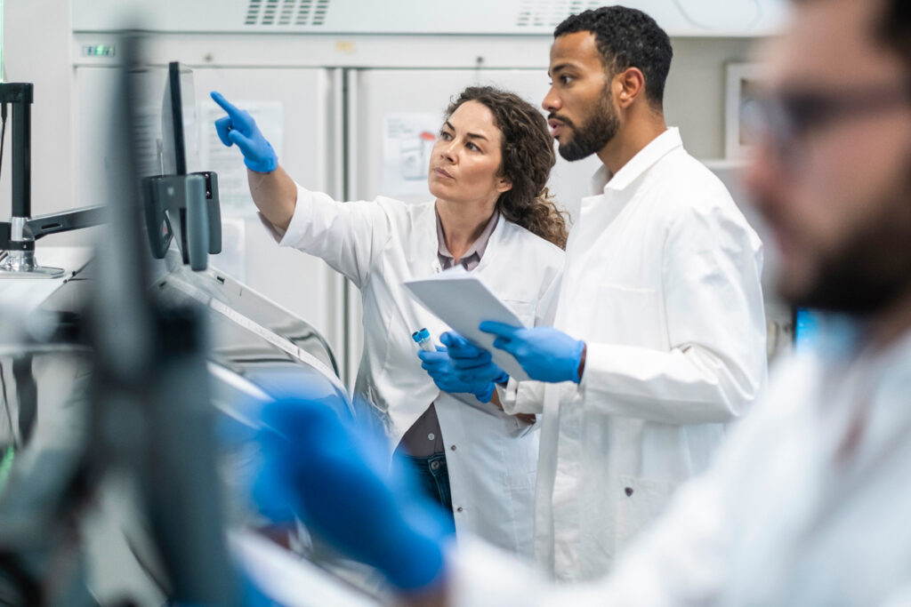 A female pharmacist in a white lab coat and blue gloves points at a monitor screen while a male colleague holds a tablet and a sample vial in a pharmaceutical lab.