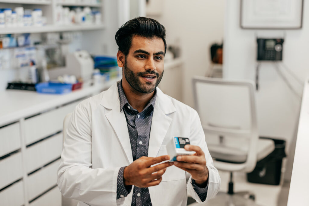 A male pharmacy technician in a white lab coat smiles while holding a medication box in a well-organized pharmacy dispensary.