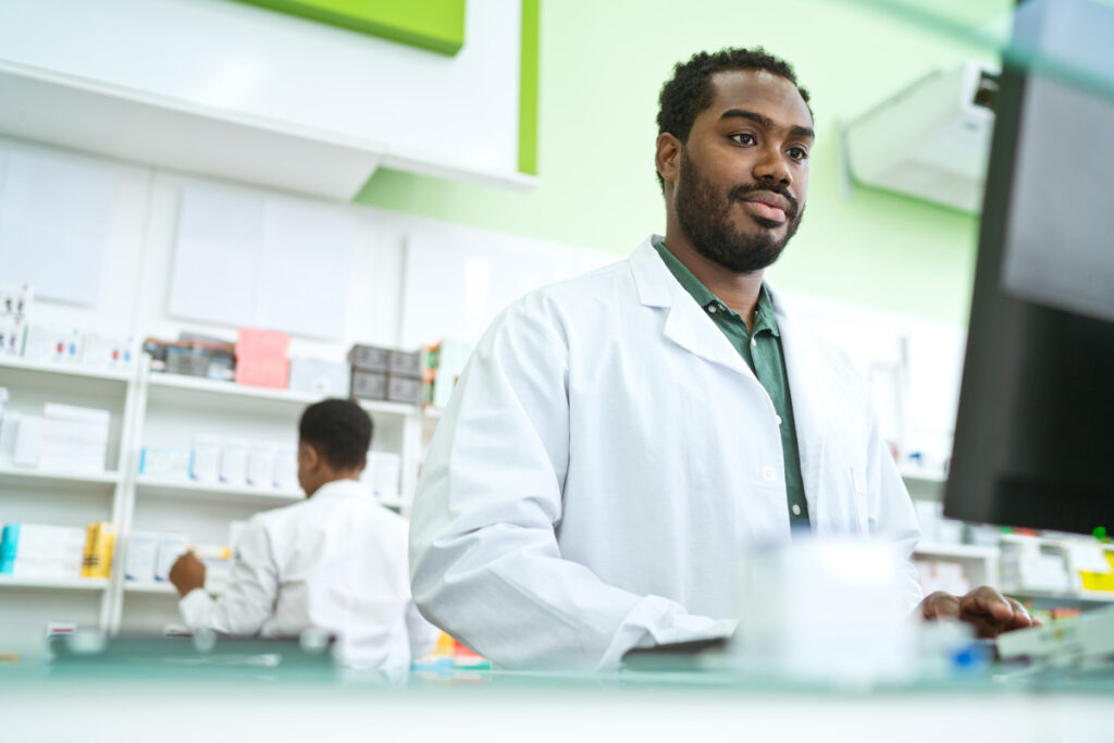 A male pharmacy technician in a white lab coat looks up from a computer at the dispensary counter, with a second technician stocking shelves in the background.