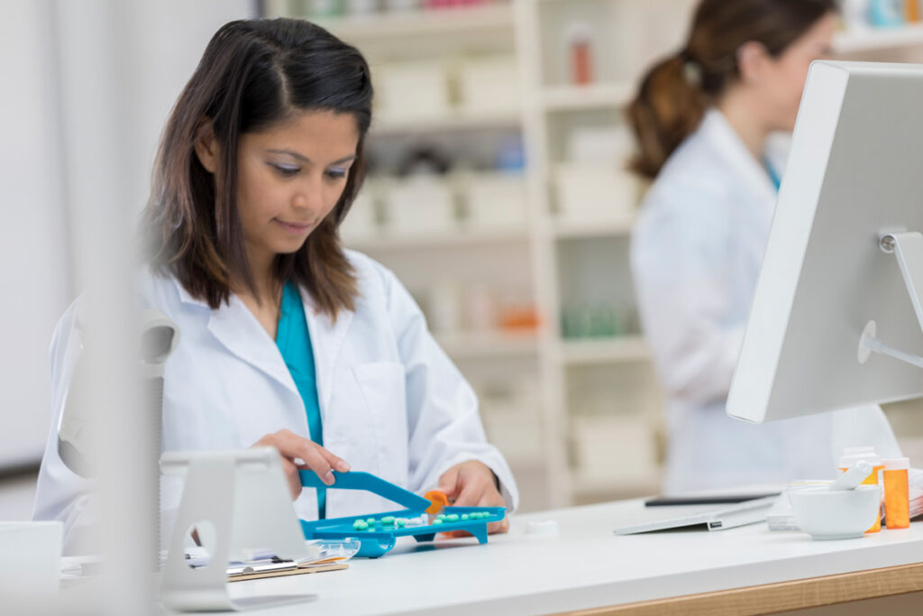 A pharmacy technician in a white lab coat uses a pill counting tray at a dispensary counter, with a colleague and computer visible in the background.