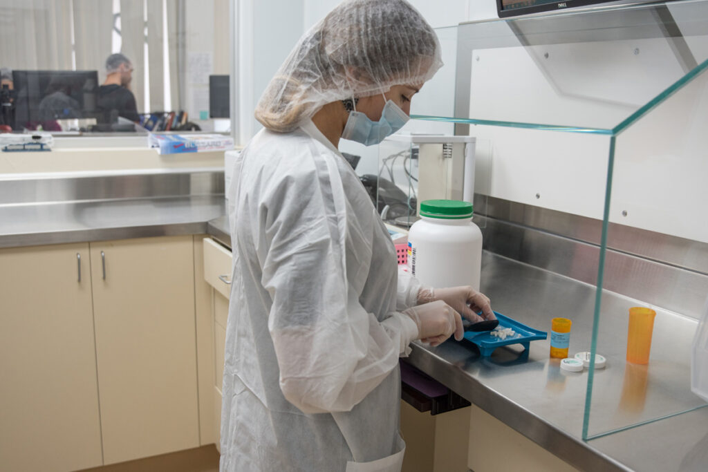 A pharmacy technician wearing a hairnet, mask, and gloves counts tablets on a dispensing tray at a compounding workstation.