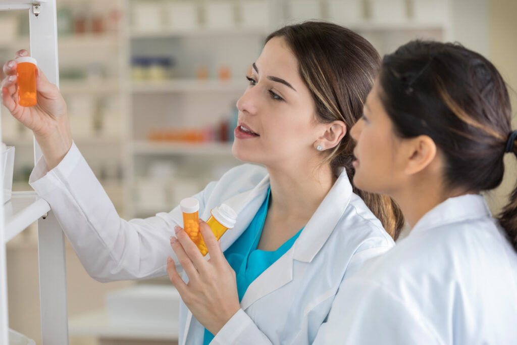 Two pharmacy technicians in white lab coats review prescription bottles together while retrieving medication from a pharmacy shelf.