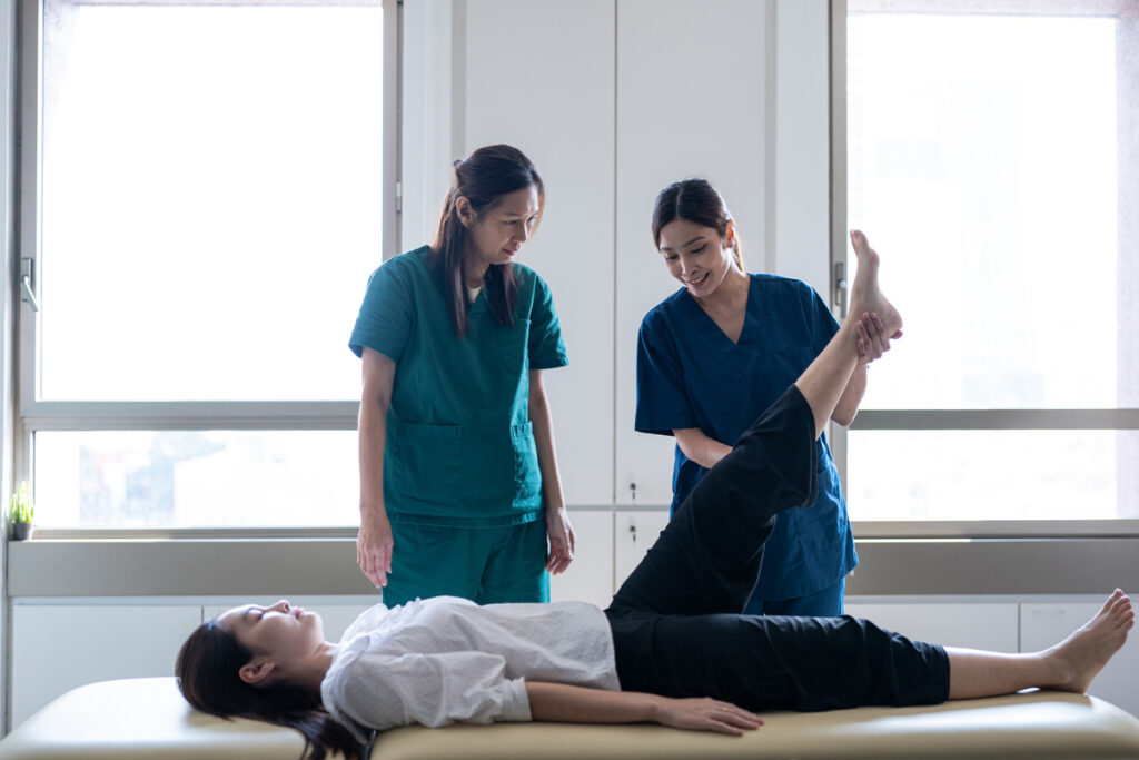 A rehabilitation assistant in blue scrubs performs a passive leg lift on a patient lying on a treatment table, observed by a supervising therapist in teal scrubs.