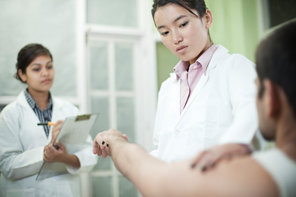 A rehabilitation assistant in a white lab coat assesses a seated male patient's wrist while a second student in a white coat takes notes on a clipboard.