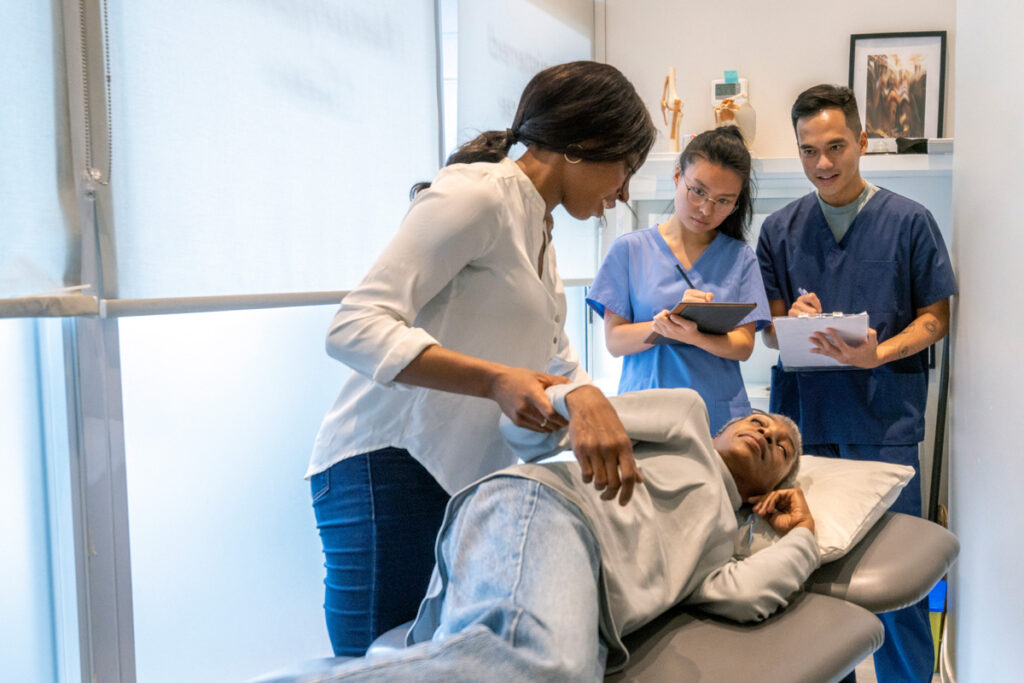 A rehabilitation instructor in a white blouse demonstrates a patient transfer technique on a treatment table while two students in scrubs observe and take notes.
