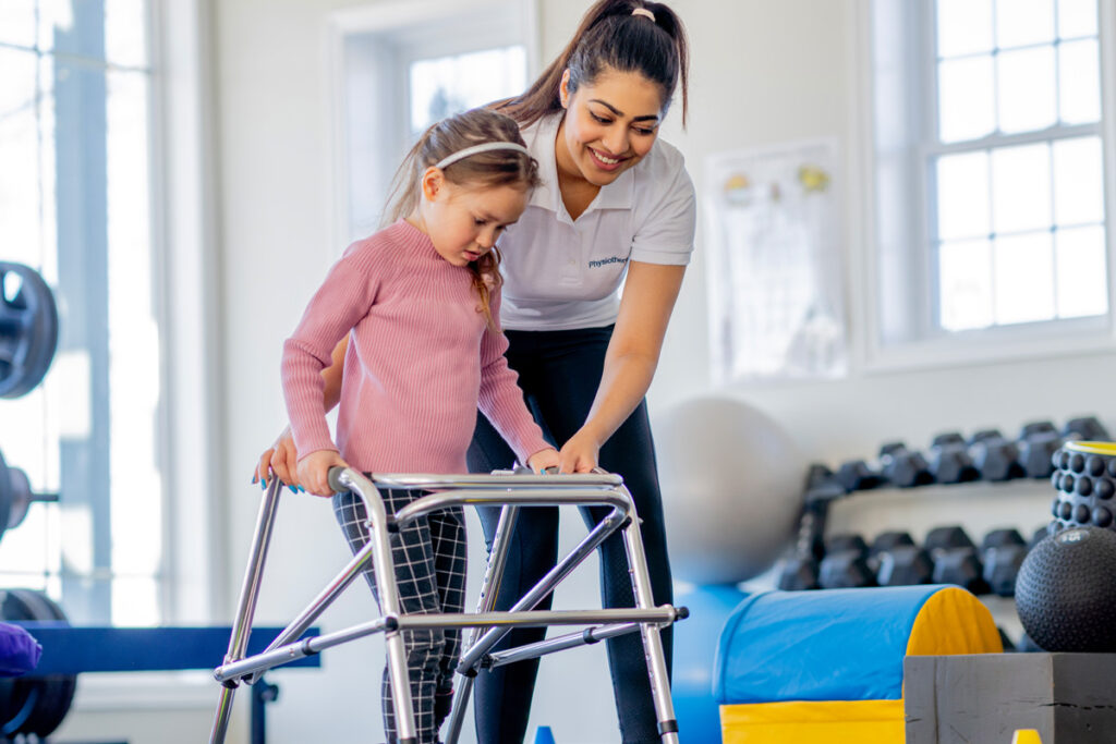 A smiling physiotherapist in a white polo shirt guides a young child using a pediatric walker during a rehabilitation session in a bright, well-equipped therapy gym.