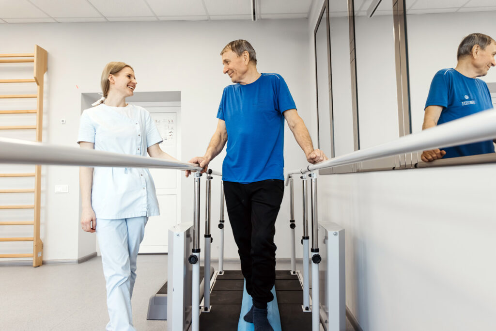 A rehabilitation assistant in light blue scrubs walks alongside an older male patient as he practices using parallel bars in a physiotherapy gym, with a mirror reflecting their progress.