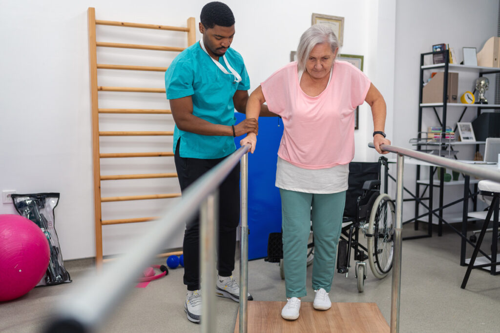 A male rehabilitation assistant in teal scrubs supports an elderly woman as she walks between parallel bars in a physiotherapy clinic, with a wheelchair visible in the background.