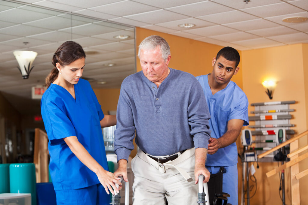 Two rehabilitation assistants in blue scrubs help an older male patient walk with a wheeled walker in a rehabilitation facility, each supporting him on either side.
