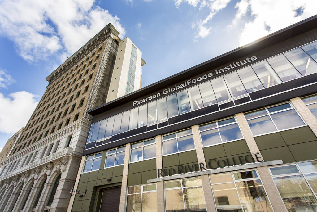 Exterior view looking up at the Paterson GlobalFoods Institute building, part of RRC Polytech, with a glass and steel modern facade attached to a historic stone tower against a partly cloudy sky.