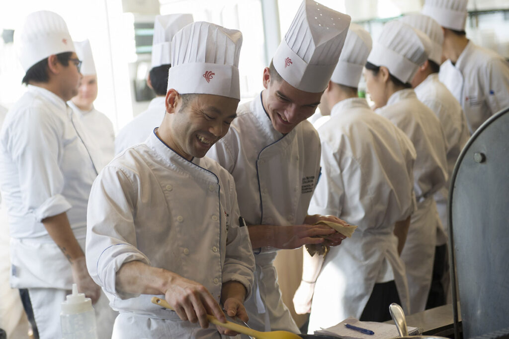 Two male culinary students in white uniforms laugh together while cooking at a stovetop in a bright, busy teaching kitchen filled with other students in the background.