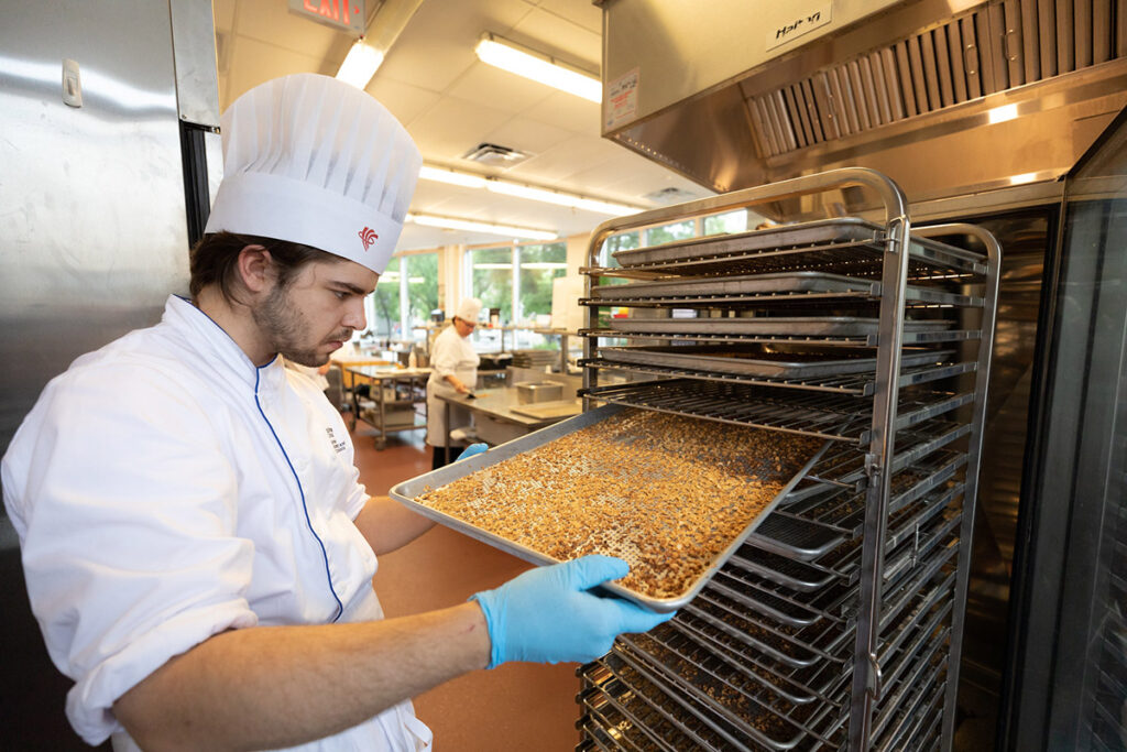 A culinary student in a white chef's coat and tall toque uses blue gloves to load a sheet pan of granola or mixed grains onto a large stainless steel rack in a professional kitchen.