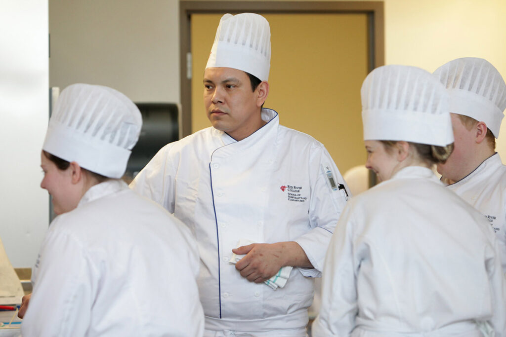 A culinary student wearing a RRC Polytech "School of Hospitality and Culinary Arts" jacket stands among a group of students in white chef uniforms and toques in a kitchen classroom.