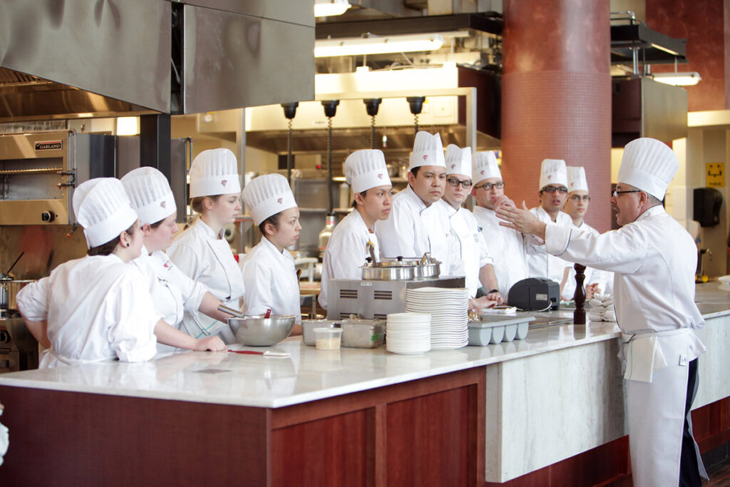 A chef instructor gestures while addressing approximately ten culinary students in white uniforms gathered around a marble-topped island in a large professional teaching kitchen.