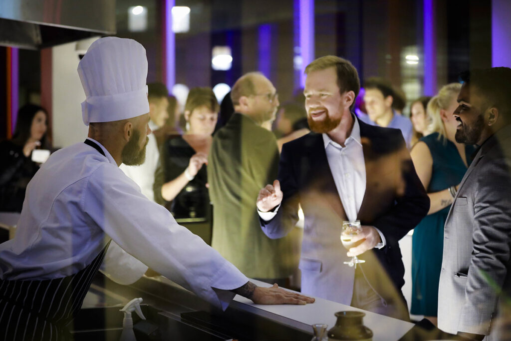A chef in a white coat and chef hat stands behind a counter conversing with two formally dressed guests at an evening networking or industry event in a warmly lit kitchen venue.