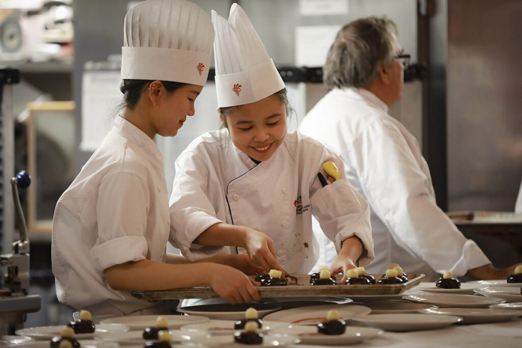 Two smiling female culinary students in white chef coats and toques carefully place chocolate and white chocolate decorations onto dessert plates, with an instructor visible in the background.