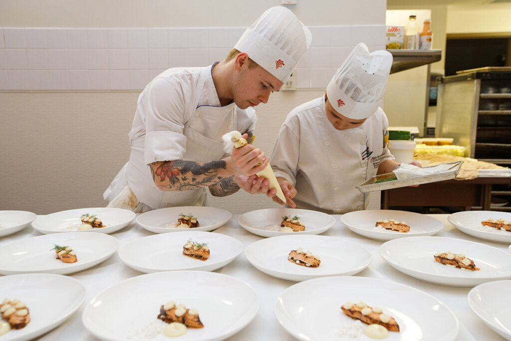 Two culinary students in white chef uniforms pipe a cream or mousse garnish onto rows of identically plated salmon dishes in a professional kitchen.