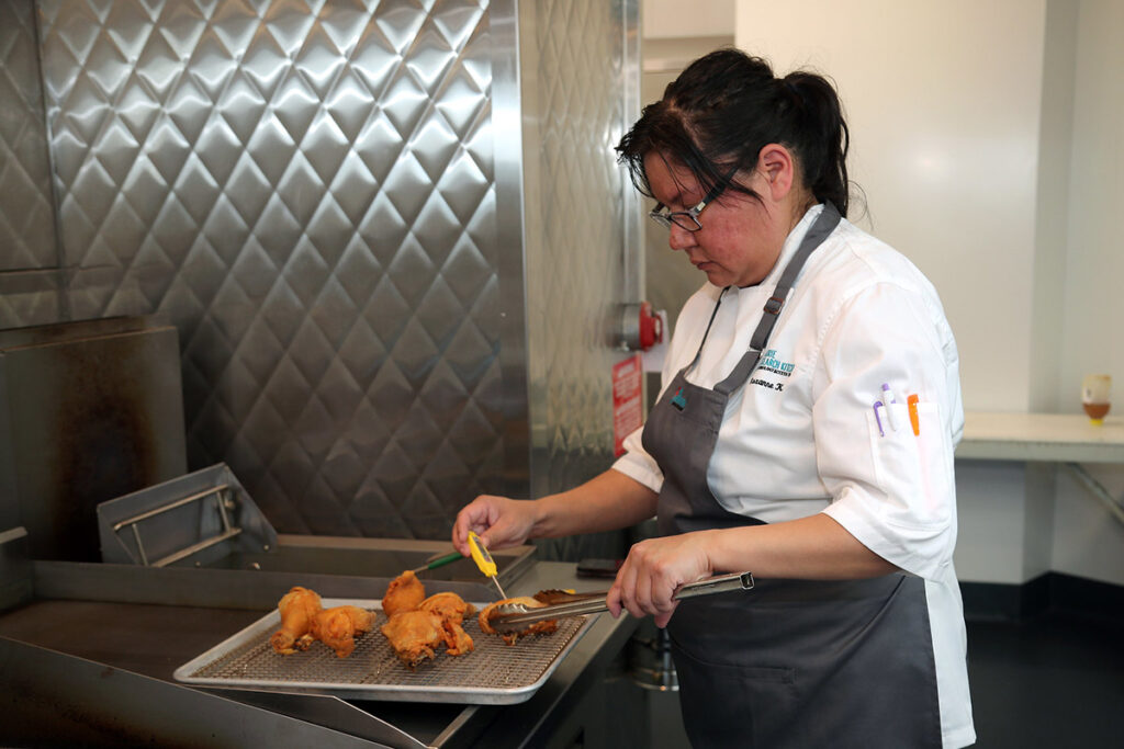 A female culinary student wearing glasses and a grey apron uses tongs and a thermometer to check the temperature of fried chicken pieces on a wire rack beside a commercial fryer.