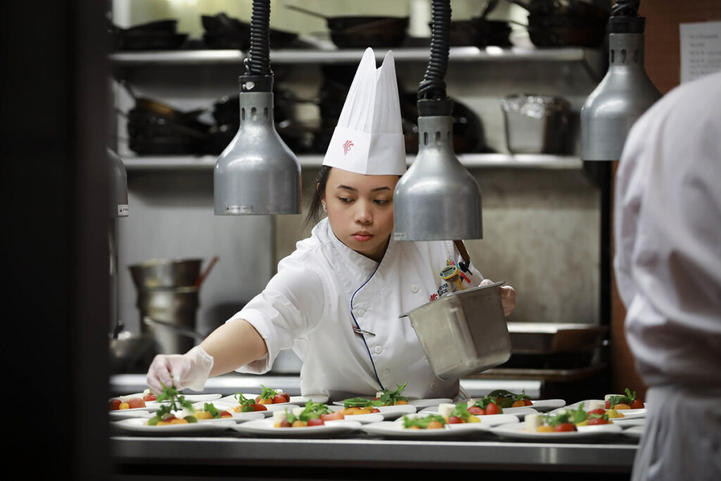 A focused female culinary student in a Red River College chef coat carefully garnishes rows of caprese-style appetizers on white spoons under heat lamps at a kitchen pass.