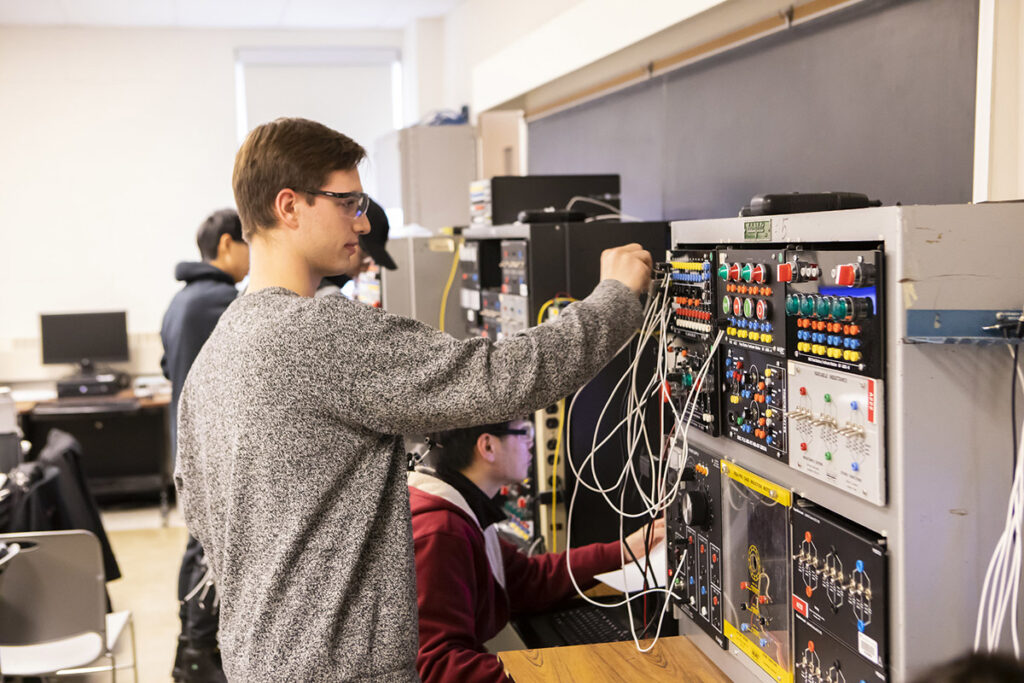 A male student in safety glasses adjusts wiring on a large electronics training panel with colourful buttons, switches, and cables, while another student works in the background.
