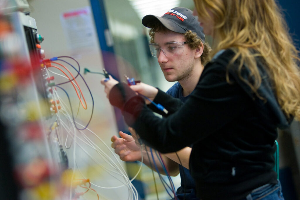A male student in a cap and safety glasses carefully connects wires on a colourful electronics training board, with a female student working beside him.