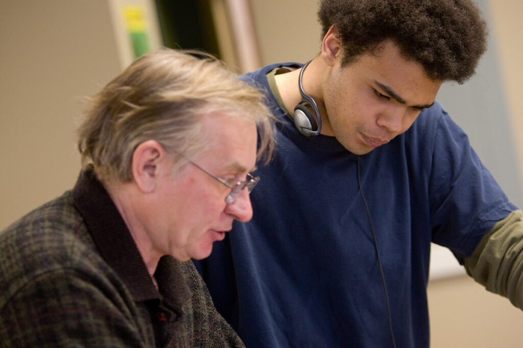 An older instructor and a younger student wearing headphones around his neck look down together at something on a desk.