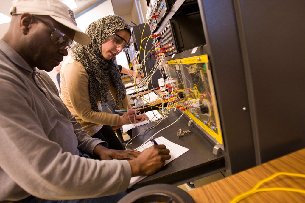 Two students — one wearing a cap and the other wearing a hijab and safety glasses — collaborate on wiring a complex electronics training panel, both taking notes on paper.