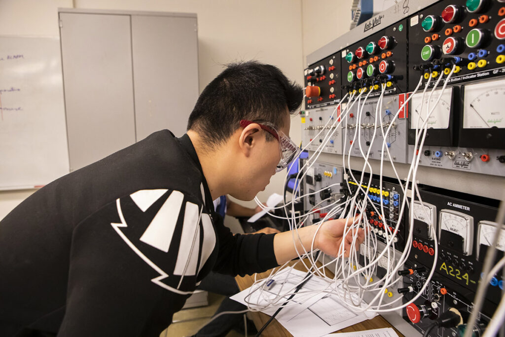 A male student wearing safety glasses leans in closely to connect wires on a Lab-Volt electronics training panel with colourful buttons, switches, and an AC ammeter, with a whiteboard visible behind him.