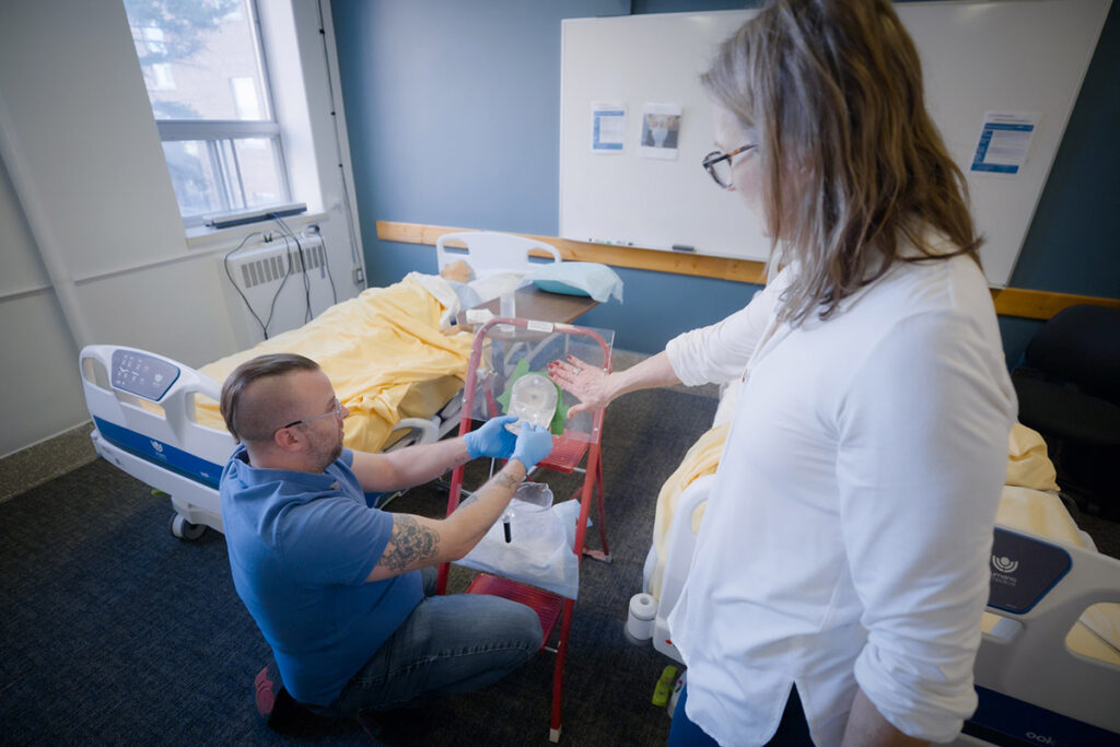 A male student in blue scrubs and gloves kneels beside a patient care aid stand holding a drainage bag, while a female instructor in a white lab coat supervises and guides him in a healthcare simulation lab.