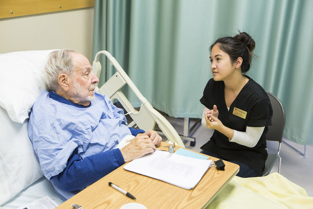 A female health care student wearing a RRC Polytech name tag sits beside an elderly male patient in a hospital bed, engaging him in a caring and attentive conversation, with a tray table and documents between them.