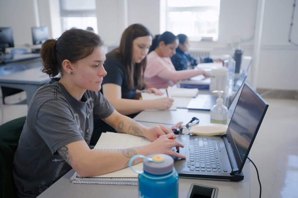 Four female students sit in a row at desks working on laptops and taking notes in a bright classroom setting, with hand sanitizer and course materials visible on the desktops.