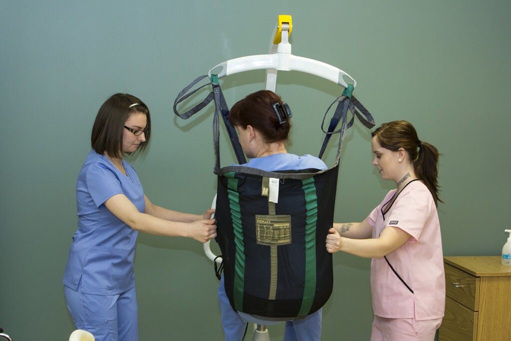 Two female health care students in scrubs — one in blue, one in pink — steady a classmate suspended in a full-body mechanical patient lift sling, practicing safe transfer techniques in a clinical simulation space.