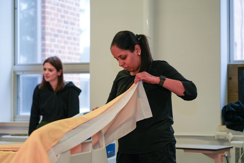 A female student in a black long-sleeve shirt carefully folds or unfolds a bed sheet over a hospital simulation bed during a bed-making skills practice, with another student visible working in the background.