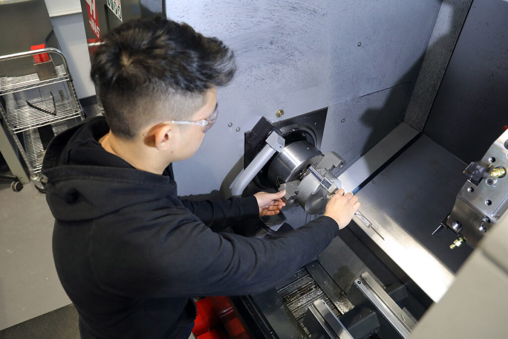 A male student wearing safety glasses uses both hands to adjust a component inside the chuck of a large Haas CNC lathe machine in an industrial machine shop lab.