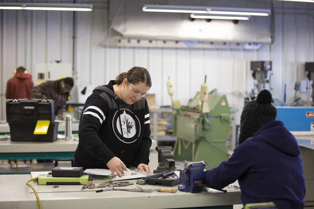 A female student in a hoodie and safety glasses works at a workshop bench examining small metal components and tools, with another student and heavy machinery visible in the background of a large industrial shop.