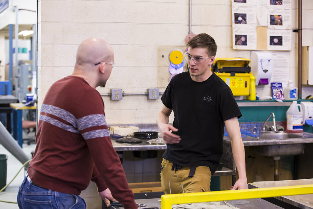 Two male students in safety glasses face each other in conversation beside a workbench in an industrial millwright lab, with instructional posters and yellow safety equipment visible on the wall behind them.