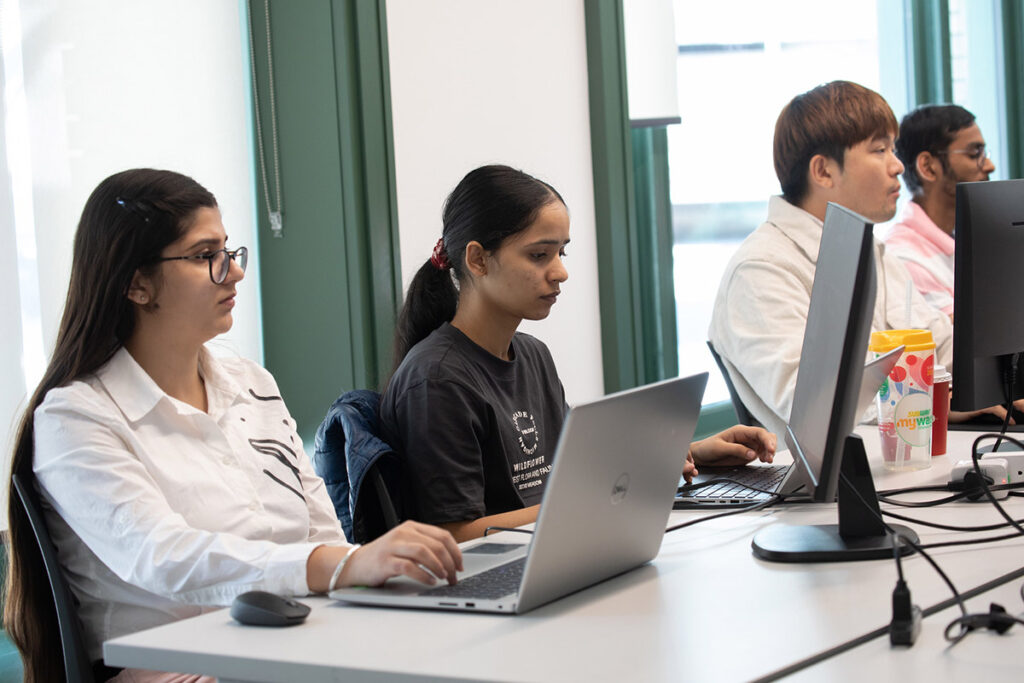 Four students — two women in the foreground and two men behind them — work independently on laptops and desktop computers in a bright, modern computer lab with green-framed windows