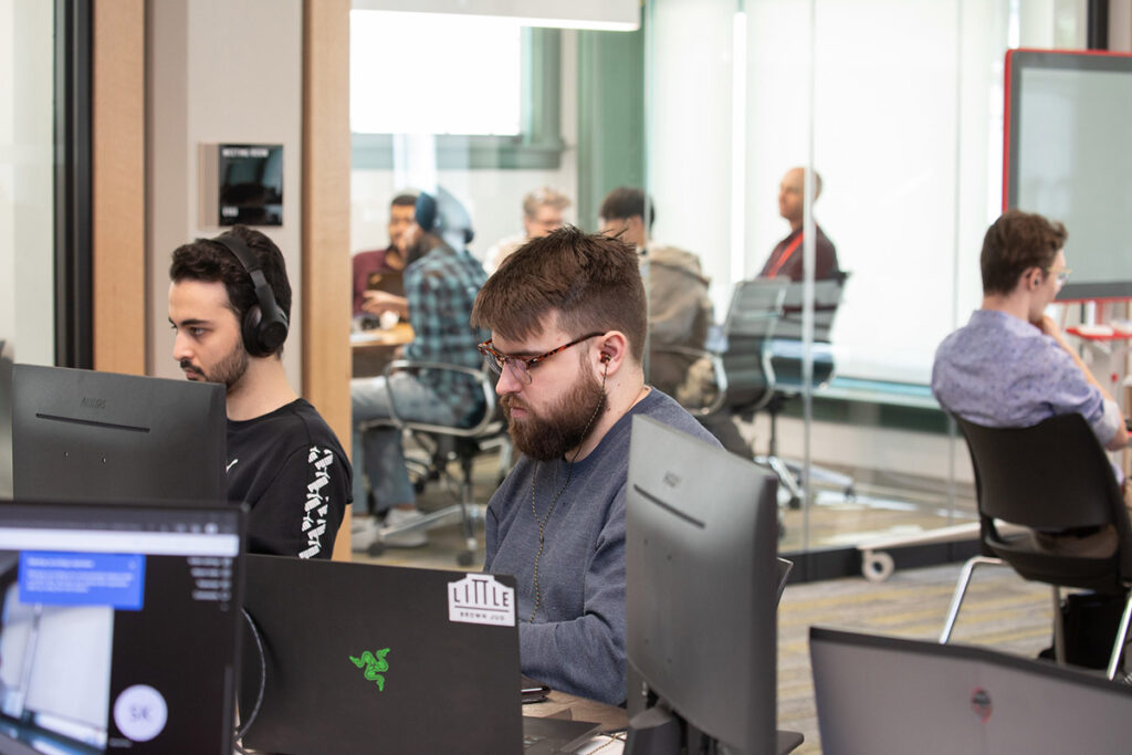 Two male students work at desktop computers in the foreground of a modern open-plan lab, with one wearing headphones and the other wearing glasses and earbuds; a glass-walled meeting room with more students is visible in the background.