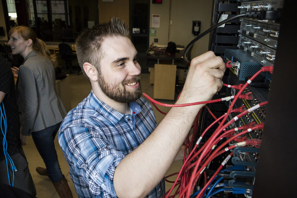 A smiling male student in a plaid shirt plugs a red ethernet cable into a rack-mounted network switch filled with bundled red, blue, and yellow cables, with a female classmate and networking lab equipment visible in the background.