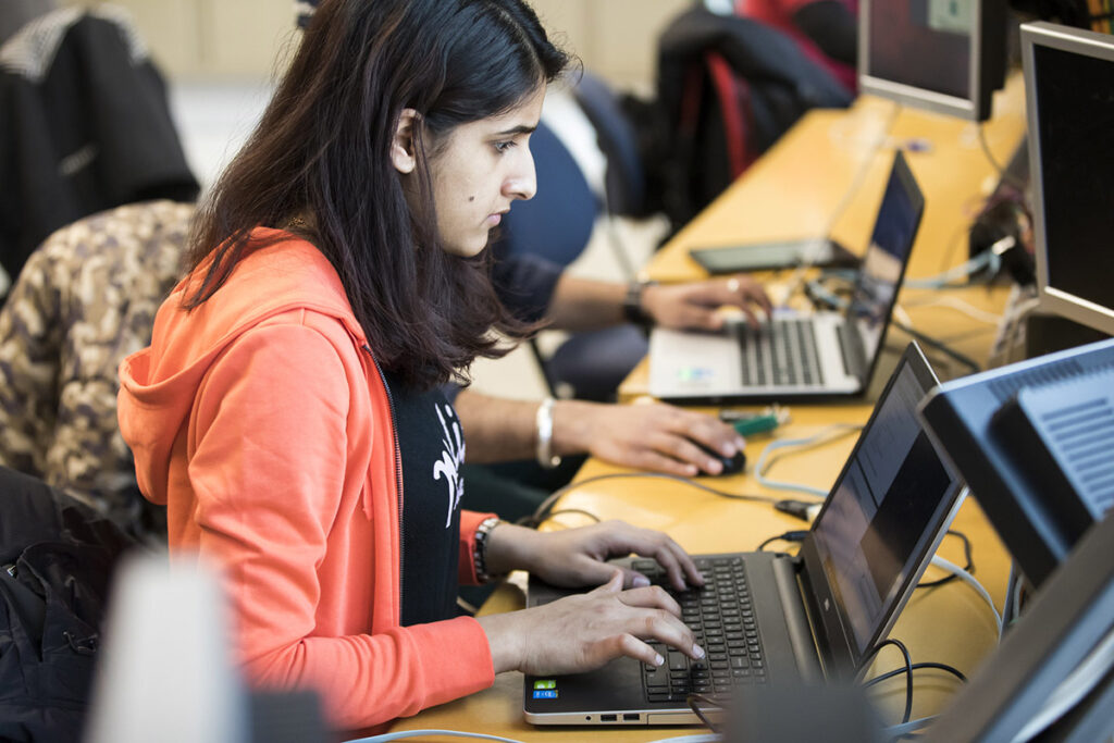 A female student in an orange hoodie types focused on a laptop in a computer lab, with other students working on laptops at a long desk beside behind her.