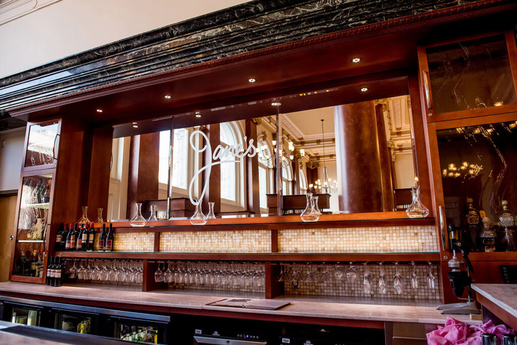 Interior of Jane's restaurant bar featuring warm wood paneling, hanging wine glasses, wine bottles, decanters, and a mirrored back bar with "Jane's" written in cursive script.