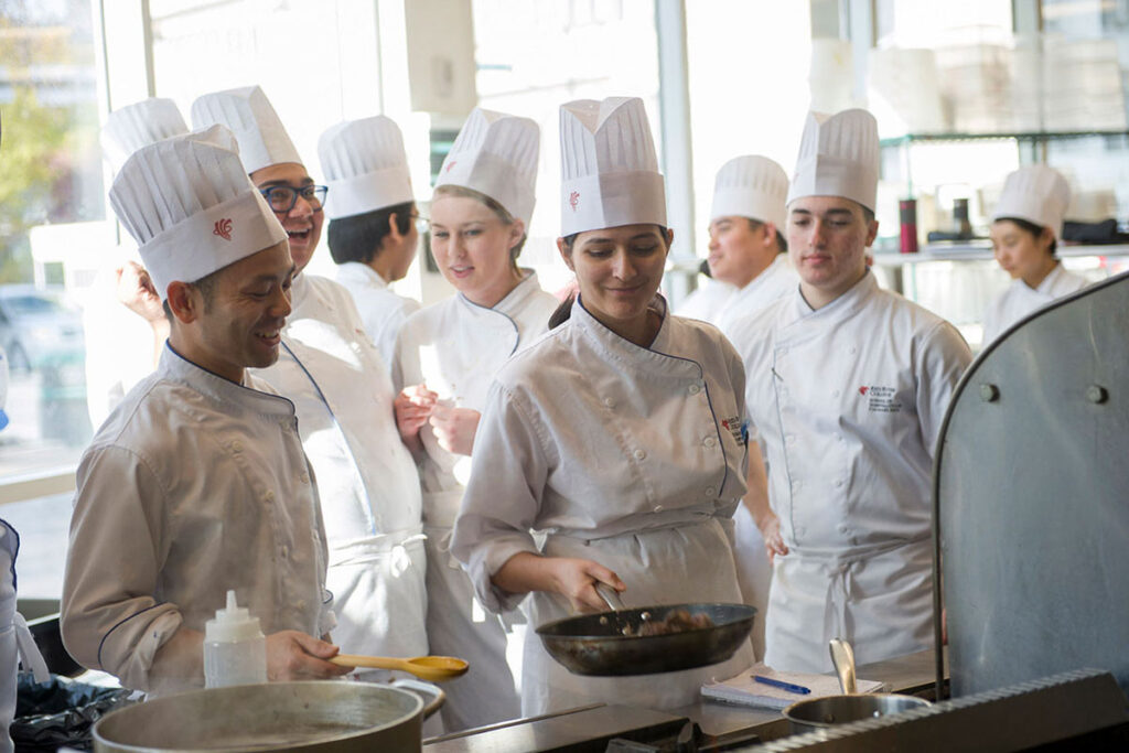 A group of culinary students in white chef coats and tall toques stand together laughing and smiling in a bright kitchen, with one student holding a wok and another holding a squeeze bottle.