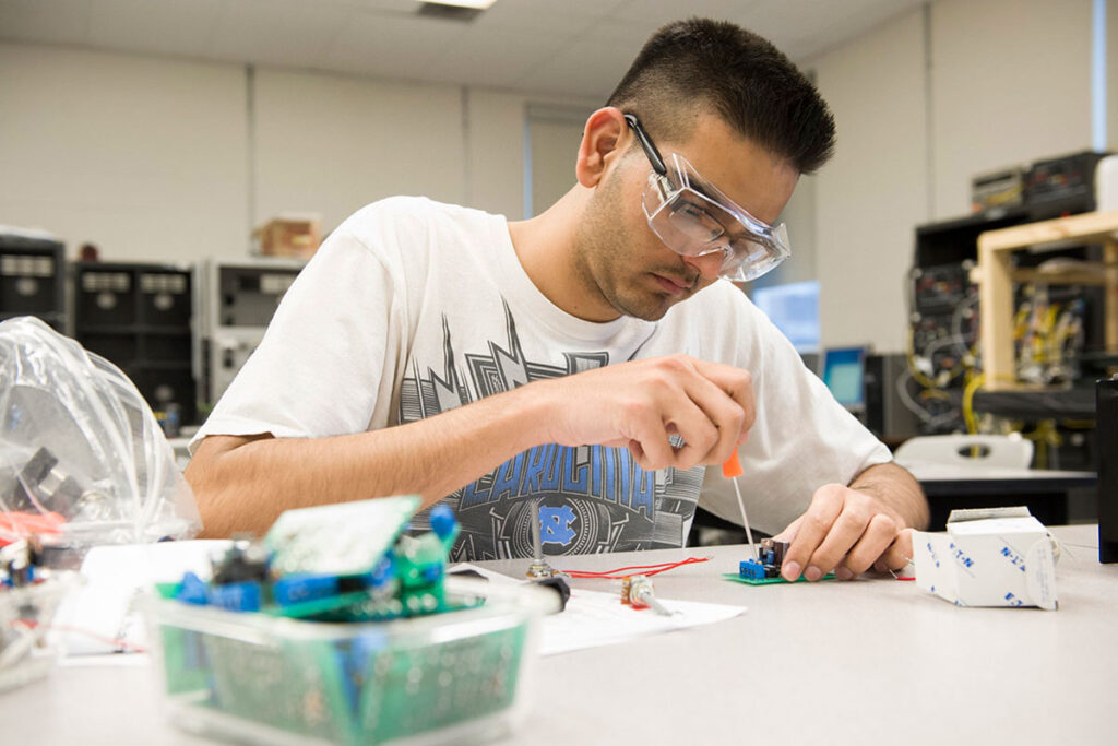 A male student wearing safety glasses and a t-shirt uses a screwdriver to work on a small circuit board at a lab bench surrounded by electronic components and equipment.